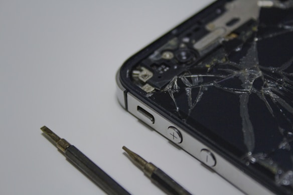 A broken smartphone with a cracked screen is placed on a surface, alongside a couple of small screwdrivers. The phone is partially disassembled and the internal components are visible.