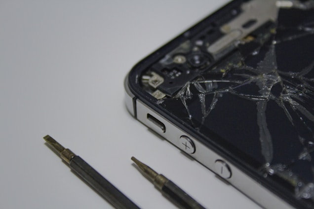 A broken smartphone with a cracked screen is placed on a surface, alongside a couple of small screwdrivers. The phone is partially disassembled and the internal components are visible.