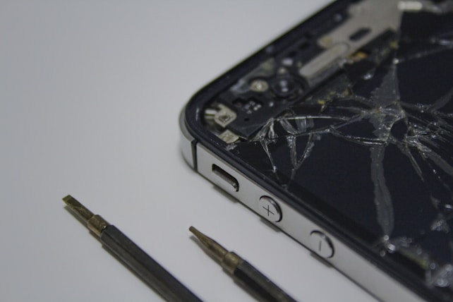 A broken smartphone with a cracked screen is placed on a surface, alongside a couple of small screwdrivers. The phone is partially disassembled and the internal components are visible.