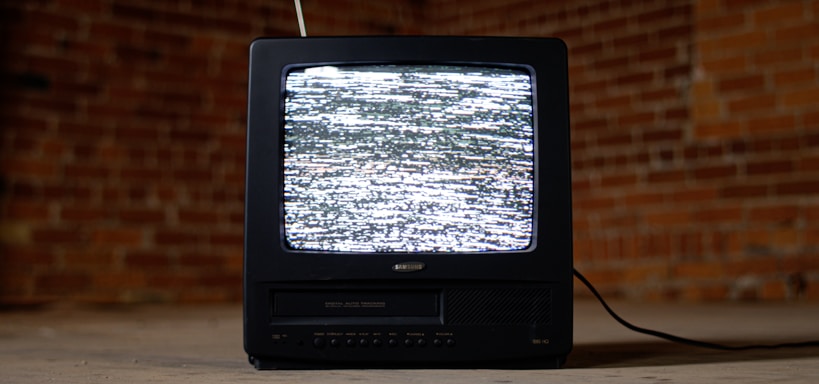 A vintage television set with a black casing is displaying static on its screen. It is placed on a concrete floor against a backdrop of red brick walls. An antenna protrudes from the top of the television, and a cable is trailing behind it on the ground.