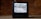 A vintage television set with a black casing is displaying static on its screen. It is placed on a concrete floor against a backdrop of red brick walls. An antenna protrudes from the top of the television, and a cable is trailing behind it on the ground.