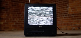 A vintage television set with a black casing is displaying static on its screen. It is placed on a concrete floor against a backdrop of red brick walls. An antenna protrudes from the top of the television, and a cable is trailing behind it on the ground.