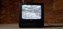 A vintage television set with a black casing is displaying static on its screen. It is placed on a concrete floor against a backdrop of red brick walls. An antenna protrudes from the top of the television, and a cable is trailing behind it on the ground.