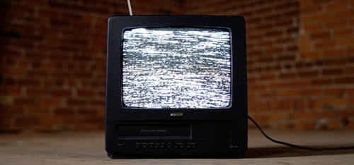 A vintage television set with a black casing is displaying static on its screen. It is placed on a concrete floor against a backdrop of red brick walls. An antenna protrudes from the top of the television, and a cable is trailing behind it on the ground.