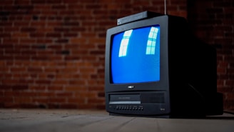 A vintage CRT television with a blue screen is placed in front of a textured brick wall. The screen reflects window panes, and the TV sits on a wooden floor, evoking a retro ambiance.