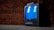 A vintage CRT television with a blue screen is placed in front of a textured brick wall. The screen reflects window panes, and the TV sits on a wooden floor, evoking a retro ambiance.