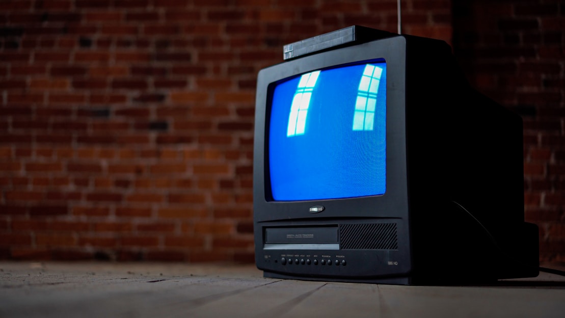 A vintage CRT television with a blue screen is placed in front of a textured brick wall. The screen reflects window panes, and the TV sits on a wooden floor, evoking a retro ambiance.
