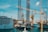 A bustling harbor scene featuring a large cruise ship docked near towering construction cranes. Various cranes and industrial equipment dominate the foreground, indicating a busy shipyard or port. The background includes a cityscape partially obscured by cranes, with calm blue waters reflecting the activities.
