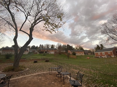A residential neighborhood with several houses surrounded by trees. The foreground features a brick patio with outdoor metal furniture, including chairs and tables. An expansive grassy backyard is visible, leading up to a row of houses. The sky is overcast with dramatic, textured clouds displaying hints of pink and orange, suggesting a sunset or sunrise.