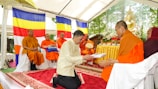 A man in a light-colored jacket is kneeling on a red carpet, offering an item wrapped in cloth to a Buddhist monk in orange robes. The scene is under a tent with Buddhist flags in the background. Several other monks are seated on chairs against the backdrop of the flags. There is an ornate golden Buddha statue and yellow floral arrangements on a table.