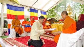 A man in a light-colored jacket is kneeling on a red carpet, offering an item wrapped in cloth to a Buddhist monk in orange robes. The scene is under a tent with Buddhist flags in the background. Several other monks are seated on chairs against the backdrop of the flags. There is an ornate golden Buddha statue and yellow floral arrangements on a table.