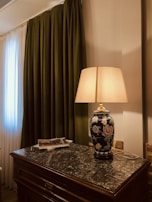 A decorative table lamp with a floral ceramic base and beige shade sits on a dark marble-topped wooden table. Next to the lamp, a stack of magazines adds a casual touch. The background features dark green floor-length curtains beside a sheer white curtain, contributing to a cozy and elegant atmosphere.