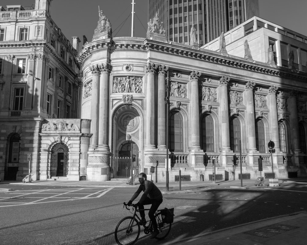 grayscale photo of man riding bicycle on road near building