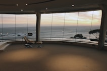 A spacious room with a large curved glass wall providing a panoramic view of the ocean and a vibrant sunset. There is a modern reclining chair with a footrest positioned near the window, and the floor is covered with a neutral-toned carpet. Outside, the water is calm, and a few small rock formations are visible. The sky displays a blend of orange, pink, and blue hues.
