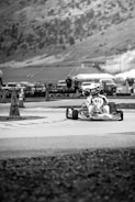 A go-kart racer navigates a paved track with traffic cones nearby. The background features a hillside with scattered trees and a few parked vehicles.
