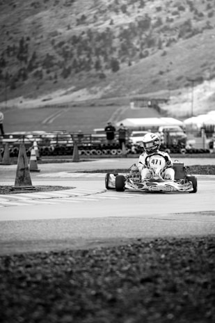 A go-kart racer navigates a paved track with traffic cones nearby. The background features a hillside with scattered trees and a few parked vehicles.