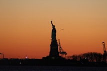 A silhouette of the Statue of Liberty stands prominently against a captivating sunset sky with hues of orange and yellow. In the background, industrial cranes and trees are faintly visible, while the calm water reflects the diminishing light. A small airplane appears in the sky, adding a sense of scale and motion.