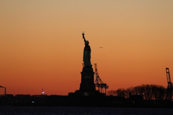 A silhouette of the Statue of Liberty stands prominently against a captivating sunset sky with hues of orange and yellow. In the background, industrial cranes and trees are faintly visible, while the calm water reflects the diminishing light. A small airplane appears in the sky, adding a sense of scale and motion.