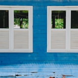Two windows with white frames and slatted blinds are set into a blue wooden wall. The paint on the wall appears weathered, with signs of peeling at the bottom. The reflection in the windows shows a green, leafy environment outside.