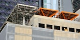 The image captures a construction site with scaffolding and exposed structural elements. Partially completed building floors are visible with metal beams and concrete surfaces. Bright orange structural frames stand out prominently. Surrounding buildings feature glass and steel facades.