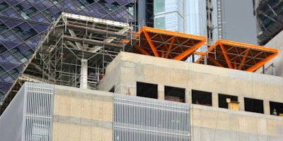 The image captures a construction site with scaffolding and exposed structural elements. Partially completed building floors are visible with metal beams and concrete surfaces. Bright orange structural frames stand out prominently. Surrounding buildings feature glass and steel facades.