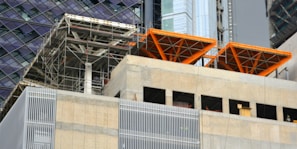 The image captures a construction site with scaffolding and exposed structural elements. Partially completed building floors are visible with metal beams and concrete surfaces. Bright orange structural frames stand out prominently. Surrounding buildings feature glass and steel facades.