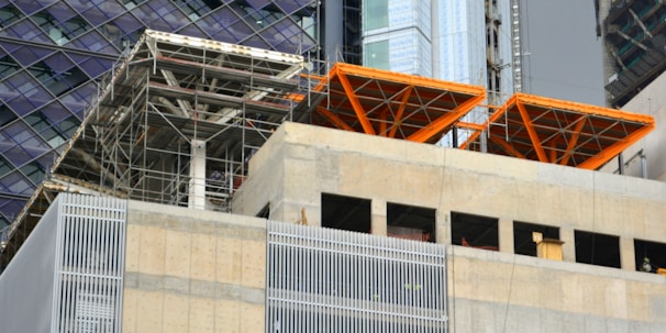 The image captures a construction site with scaffolding and exposed structural elements. Partially completed building floors are visible with metal beams and concrete surfaces. Bright orange structural frames stand out prominently. Surrounding buildings feature glass and steel facades.