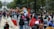 A vibrant outdoor festival with people walking along a street, surrounded by colorful parade floats and decorations. Trees line the path and a prominent fountain with a statue is visible in the background. Participants carry red flags and there is a lively atmosphere.