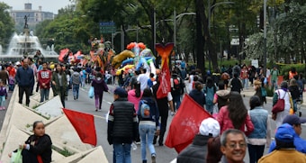 A vibrant outdoor festival with people walking along a street, surrounded by colorful parade floats and decorations. Trees line the path and a prominent fountain with a statue is visible in the background. Participants carry red flags and there is a lively atmosphere.