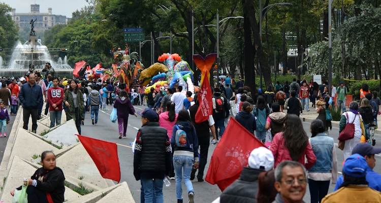A vibrant outdoor festival with people walking along a street, surrounded by colorful parade floats and decorations. Trees line the path and a prominent fountain with a statue is visible in the background. Participants carry red flags and there is a lively atmosphere.