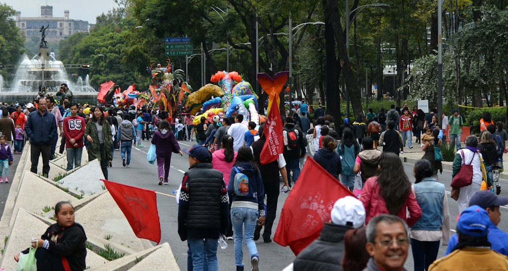 A vibrant outdoor festival with people walking along a street, surrounded by colorful parade floats and decorations. Trees line the path and a prominent fountain with a statue is visible in the background. Participants carry red flags and there is a lively atmosphere.