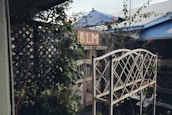 A rustic garden scene featuring an ornate metal plant stand in the foreground. To the left, climbing plants on a wooden trellis create a lush, green backdrop. In the background, there is a sign with the letters 'BLM' and several blue-tinted structures partially visible.