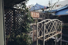 A rustic garden scene featuring an ornate metal plant stand in the foreground. To the left, climbing plants on a wooden trellis create a lush, green backdrop. In the background, there is a sign with the letters 'BLM' and several blue-tinted structures partially visible.