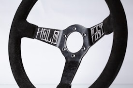 A black racing steering wheel with a minimalist and rugged design, featuring the words 'HOLD' and 'FIGHT' engraved on its metallic spokes. The wheel is set against a plain white background, enhancing the focus on its sleek and functional aesthetic.