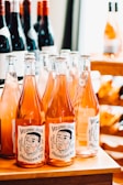 A collection of wine bottles is displayed on a wooden surface. The front row features bottles with orange-pink colored liquid, with labels reading 'Weeping Juan' and depicting a sketched face. In the background, there are other bottles with red wine, some partially visible, creating a well-stocked and organized setting.