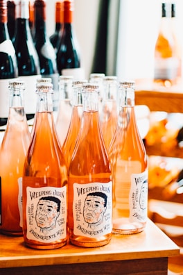 A collection of wine bottles is displayed on a wooden surface. The front row features bottles with orange-pink colored liquid, with labels reading 'Weeping Juan' and depicting a sketched face. In the background, there are other bottles with red wine, some partially visible, creating a well-stocked and organized setting.