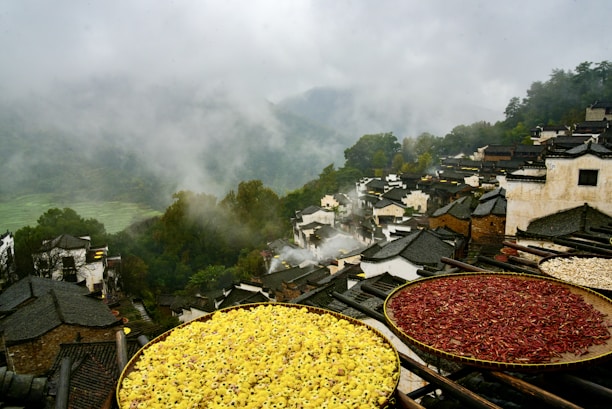 A misty mountain village with traditional dark-tiled rooftops nestled among lush green hills. Large round baskets with drying herbs and red chilies are in the foreground, adding vibrant colors to the scene. Smoke rises gently, giving a serene and timeless atmosphere.