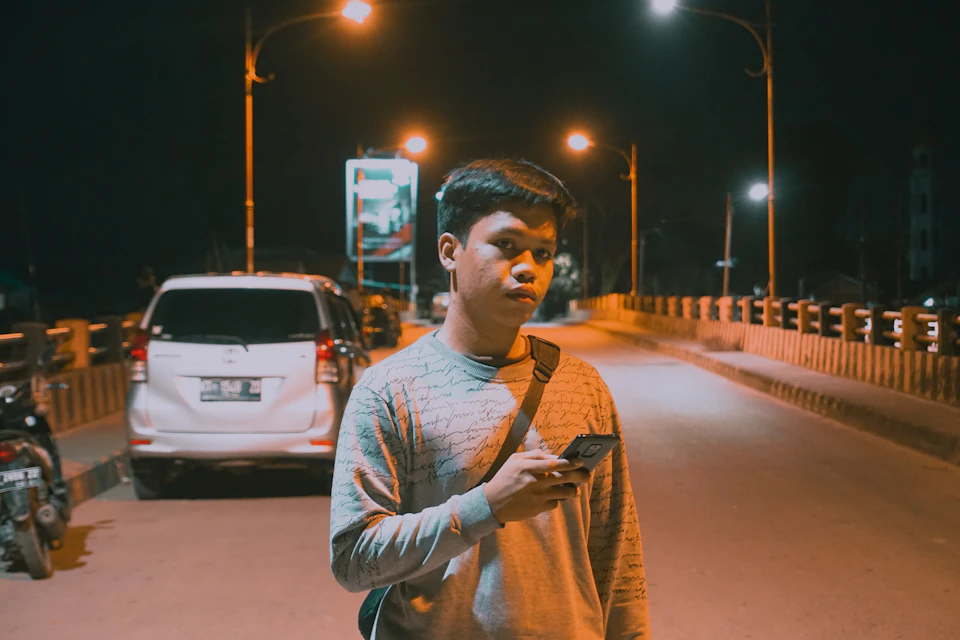 The image depicts a young man standing on a street at night, engrossed in his phone. He is casually dressed in a gray sweatshirt and has a backpack slung over one shoulder. The background features a bridge with two street lamps casting a warm glow, illuminating the scene. In the distance, there are parked cars lining the street, adding to the urban atmosphere of the image.