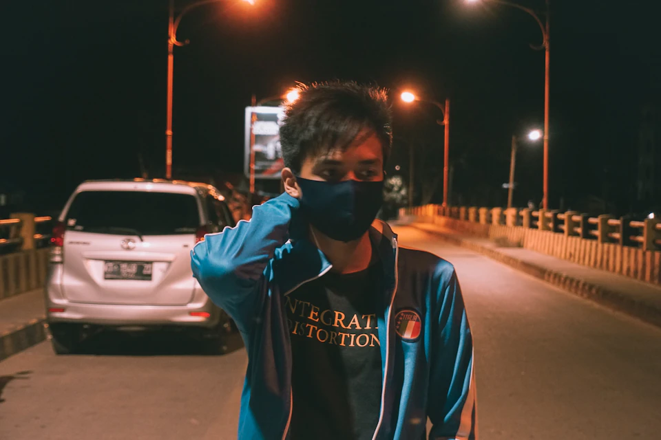 The image depicts a young man wearing a blue jacket and a face mask, standing on a bridge at night with his hand raised to his face. The bridge is illuminated by street lamps that cast a warm glow over the scene. In the background, there are two cars parked along the side of the road - one white car and one silver car.