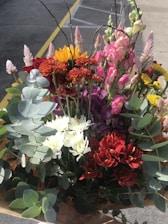 A vibrant bouquet featuring a variety of flowers, including sunflowers, red chrysanthemums, white daisies, and snapdragons. The bouquet is accented with eucalyptus leaves and other greenery. The assortment of colors and textures creates a dynamic and eye-catching arrangement.