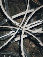 A complex network of intersecting highway overpasses captured from an aerial perspective, showcasing the intricacy and scale of modern infrastructure. Multiple lanes of traffic can be seen with vehicles of various sizes traveling on the roads. The surrounding landscape includes patches of greenery and some exposed earth.