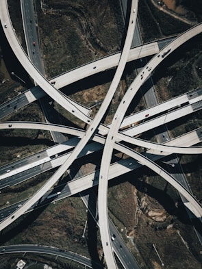 A complex network of intersecting highway overpasses captured from an aerial perspective, showcasing the intricacy and scale of modern infrastructure. Multiple lanes of traffic can be seen with vehicles of various sizes traveling on the roads. The surrounding landscape includes patches of greenery and some exposed earth.