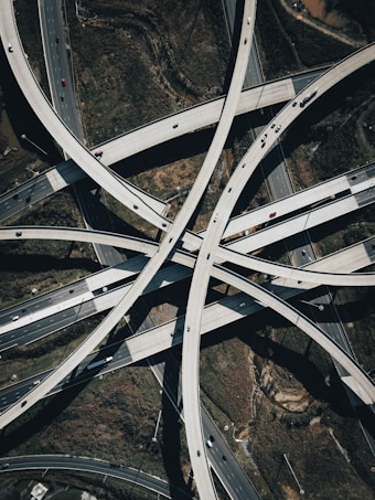 A complex network of intersecting highway overpasses captured from an aerial perspective, showcasing the intricacy and scale of modern infrastructure. Multiple lanes of traffic can be seen with vehicles of various sizes traveling on the roads. The surrounding landscape includes patches of greenery and some exposed earth.