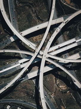 A complex network of intersecting highway overpasses captured from an aerial perspective, showcasing the intricacy and scale of modern infrastructure. Multiple lanes of traffic can be seen with vehicles of various sizes traveling on the roads. The surrounding landscape includes patches of greenery and some exposed earth.