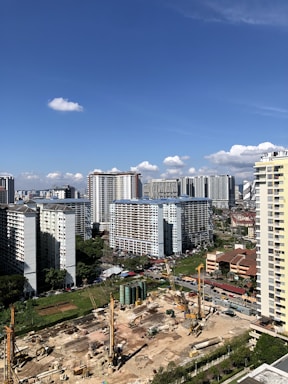 A cityscape featuring high-rise buildings in a residential area. In the foreground, there is a construction site with large machinery, including cranes and cylindrical building materials. The weather appears clear with a blue sky and a few scattered clouds.