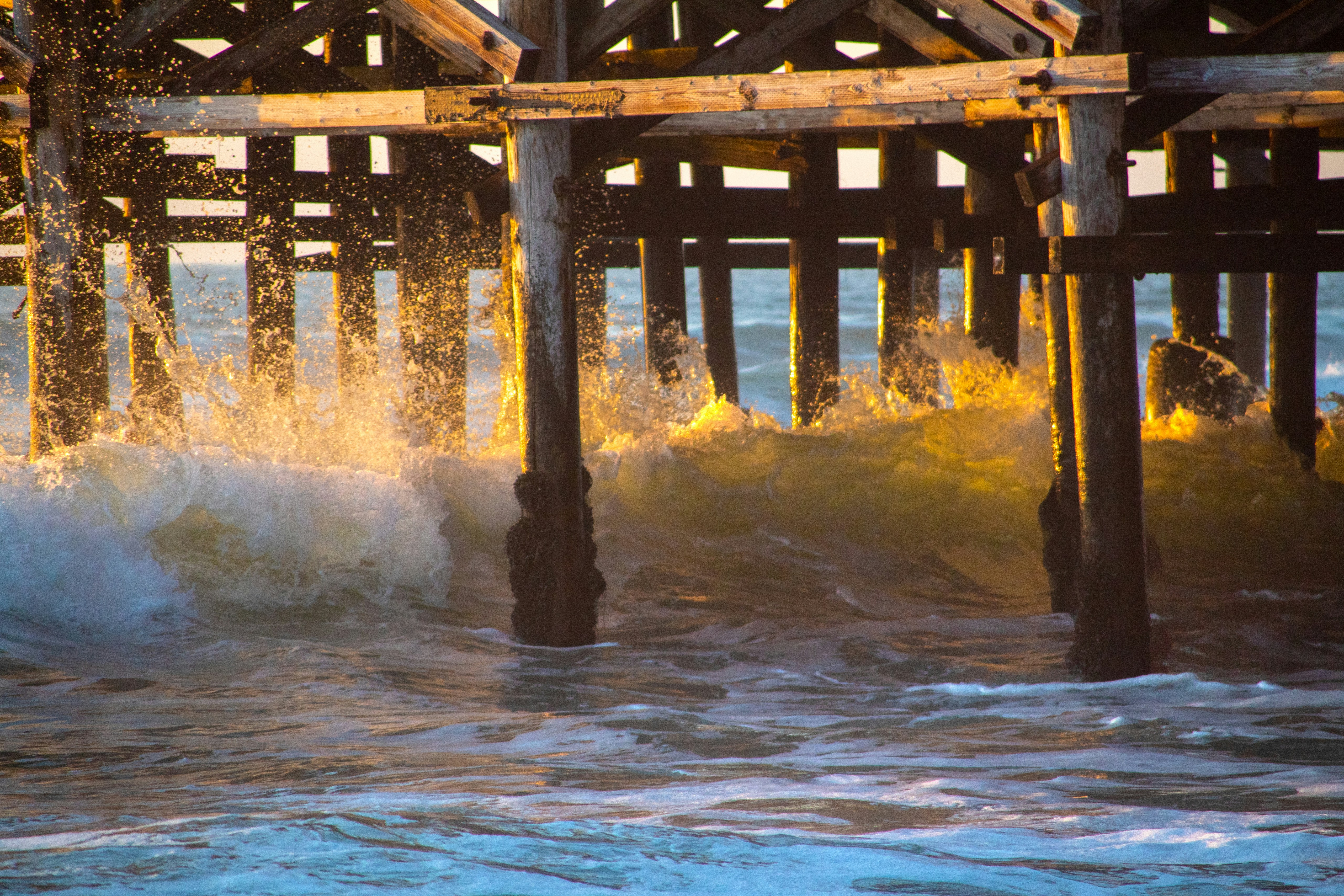brown wooden dock on water, Wave crashing into Crystal Pier in Pacific Beach, San Diego, California, with orange and blue hues.