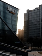 A cityscape featuring modern office buildings. The foreground displays a building with glass walls and a logo, while a taller structure in the background has the Microsoft logo. Trees and a bicycle are visible at ground level, and the sunlight is filtering through the buildings, casting a warm glow.