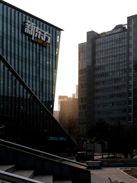 A cityscape featuring modern office buildings. The foreground displays a building with glass walls and a logo, while a taller structure in the background has the Microsoft logo. Trees and a bicycle are visible at ground level, and the sunlight is filtering through the buildings, casting a warm glow.