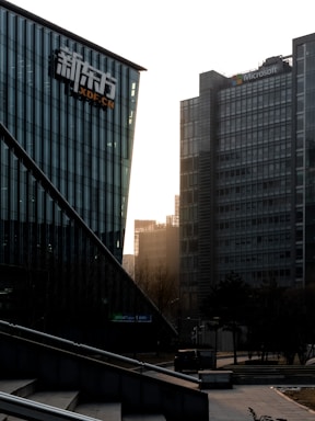 A cityscape featuring modern office buildings. The foreground displays a building with glass walls and a logo, while a taller structure in the background has the Microsoft logo. Trees and a bicycle are visible at ground level, and the sunlight is filtering through the buildings, casting a warm glow.