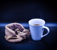 A steaming cup of coffee sits next to a burlap sack spilling over with roasted coffee beans, set against a dark background.